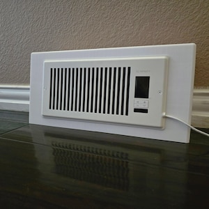 May include: A white AC Infinity vent cover with black vertical slats. The cover has a digital display and control buttons. The unit is rectangular and mounted on a white frame. A white cord extends from the side.