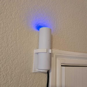 Op de afbeelding: Een wit, cilindrisch smart home-apparaat dat aan een muur is bevestigd en blauw licht van bovenaf uitstraalt. Het apparaat wordt op zijn plaats gehouden door een witte beugel en is aangesloten op een zwarte kabel.