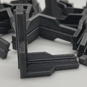 Puede incluir: Una colección de escuadras de esquina de plástico negro en forma de L. Las escuadras están apiladas y dispersas, con un enfoque en las superficies texturizadas y el diseño en ángulo recto. La imagen está bien iluminada, destacando los detalles de los componentes impresos en 3D.