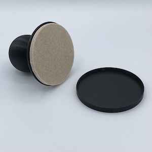Puede incluir: Un sello negro y blanco con una almohadilla redonda de fieltro beige. El sello tiene una base redonda y un mango con forma de rectángulo redondeado.