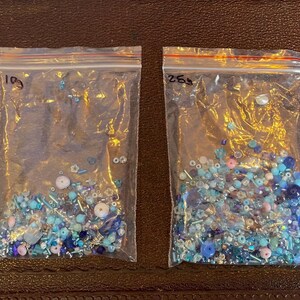 Op de afbeelding: Twee doorzichtige plastic zakken gevuld met diverse blauwe kralen. De kralen vari&euml;ren in grootte en vorm, waaronder ronde, cilindrische en onregelmatige vormen. Sommige kralen zijn iriserend, andere ondoorzichtig. De zakken zijn gelabeld met handgeschreven nummers.