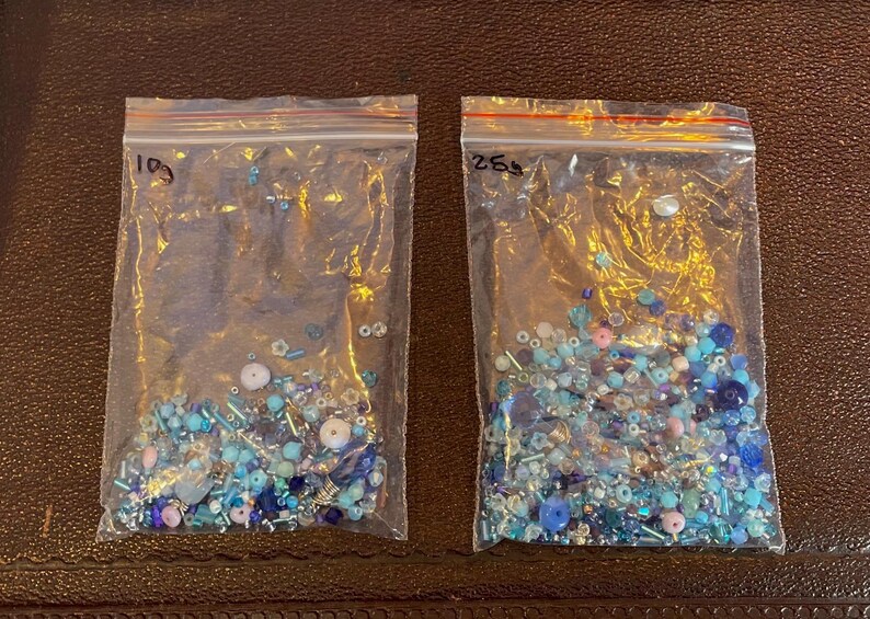Op de afbeelding: Twee doorzichtige plastic zakken gevuld met diverse blauwe kralen. De kralen vari&euml;ren in grootte en vorm, waaronder ronde, cilindrische en onregelmatige vormen. Sommige kralen zijn iriserend, andere ondoorzichtig. De zakken zijn gelabeld met handgeschreven nummers.