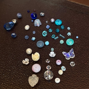 Op de afbeelding: Een verzameling kralen in verschillende tinten blauw, turquoise en wit. De collectie bevat ronde, facetgeslepen en bloemvormige kralen, plus een vlindervormige kraal. De kralen liggen op een donkerbruin oppervlak.