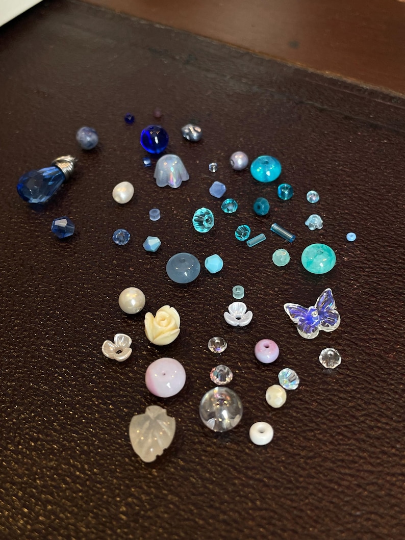 Op de afbeelding: Een verzameling kralen in verschillende tinten blauw, turquoise en wit. De collectie bevat ronde, facetgeslepen en bloemvormige kralen, plus een vlindervormige kraal. De kralen liggen op een donkerbruin oppervlak.