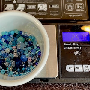 Op de afbeelding: Een witte kom gevuld met een assortiment blauwe kralen, waaronder verschillende vormen en maten, staat op een digitale weegschaal. De weegschaal geeft een gewicht van 25,70 aan. De weegschaal heeft een capaciteit van 200g en een aflezing van 0,01g.