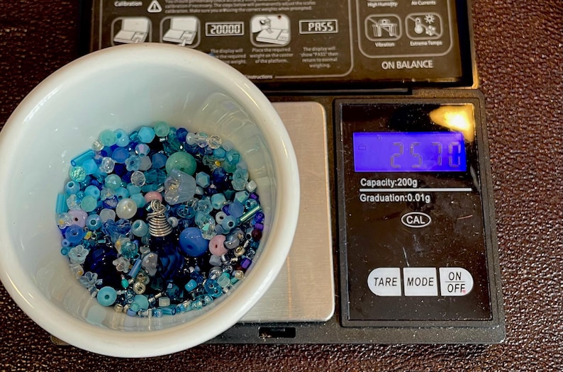Op de afbeelding: Een witte kom gevuld met een assortiment blauwe kralen, waaronder verschillende vormen en maten, staat op een digitale weegschaal. De weegschaal geeft een gewicht van 25,70 aan. De weegschaal heeft een capaciteit van 200g en een aflezing van 0,01g.