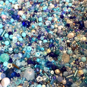 Op de afbeelding: Een close-up van een mix van blauwe kralen, vari&euml;rend van lichtblauw tot diep marineblauw. De kralen hebben verschillende vormen en maten, waaronder rond, facetgeslepen en buisvormig. Sommige kralen zijn helder, andere ondoorzichtig of iriserend.