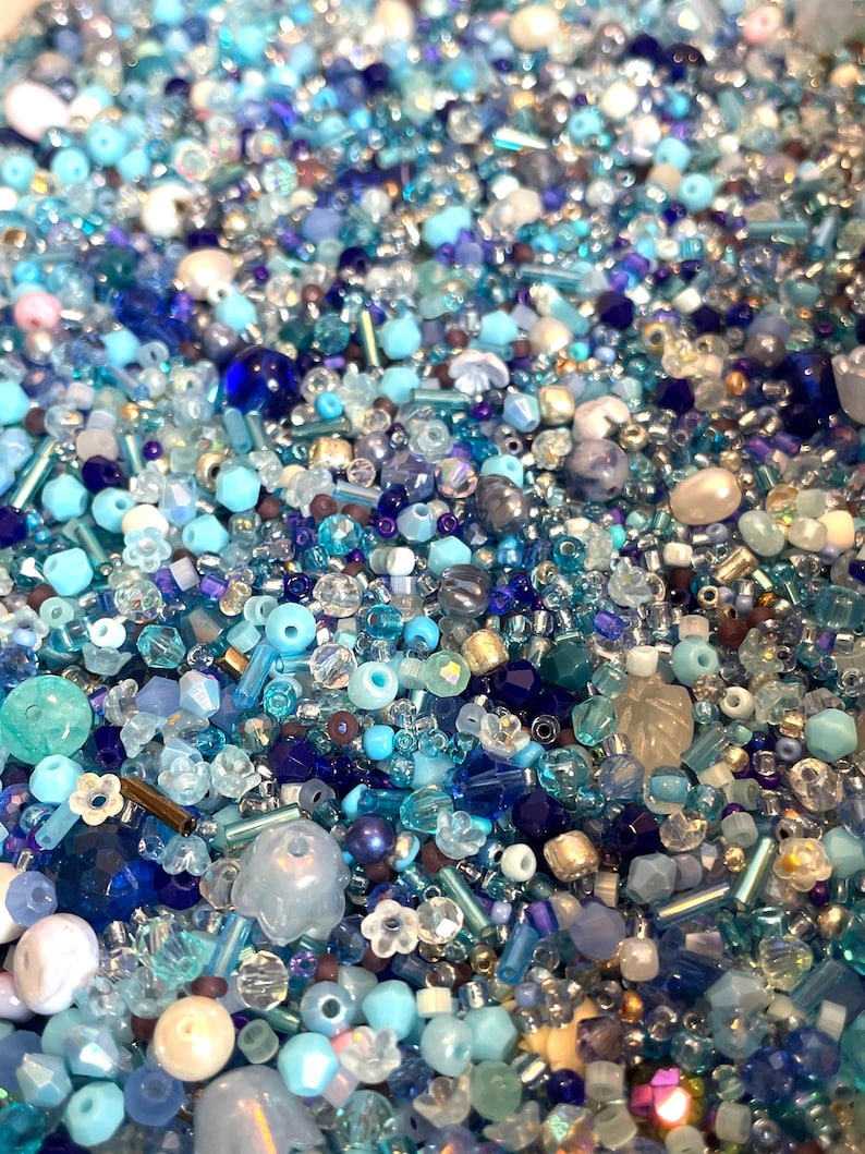 Op de afbeelding: Een close-up van een mix van blauwe kralen, vari&euml;rend van lichtblauw tot diep marineblauw. De kralen hebben verschillende vormen en maten, waaronder rond, facetgeslepen en buisvormig. Sommige kralen zijn helder, andere ondoorzichtig of iriserend.