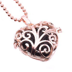 Puede incluir: Un collar con un dije en forma de corazón de oro rosa con un delicado diseño de filigrana. El dije está abierto y revela una roca de lava negra en su interior.