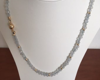 May include: A beaded necklace featuring small, faceted, light blue translucent beads interspersed with tiny, polished gold-tone metal spheres. The necklace includes a larger, round, gold-tone magnetic clasp closure positioned on the left side.