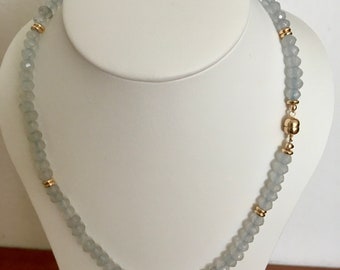 May include: A necklace featuring faceted light blue beads arranged in a circular pattern, accented with small gold-toned metal spacers at regular intervals. The piece is secured with a round gold-toned magnetic clasp positioned on the right side.