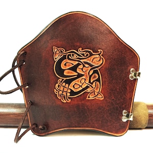 Puede incluir: Un protector de brazo de cuero marrón con un diseño de nudo celta. El protector tiene una forma curva, con un patrón decorativo en relieve en el centro. Cuenta con cordones de cuero y cierres metálicos.