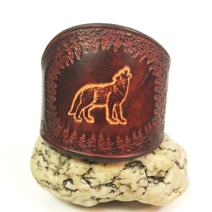 May include: A dark brown leather cuff bracelet with a howling wolf design. The wolf is etched in the center, surrounded by a forest border. The bracelet is approximately 2 inches wide.