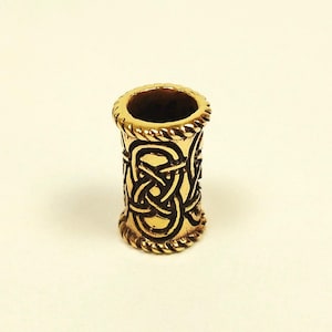 May include: A gold-colored metal bead with a Celtic knot design. The bead is cylindrical with a rope-like texture at the top and bottom. The Celtic knot pattern is dark brown against the gold background.
