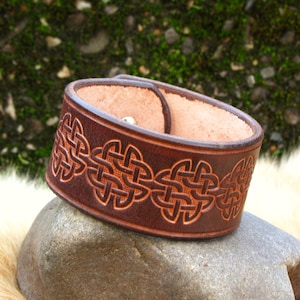 May include: A brown leather bracelet with a Celtic knot design. The bracelet is wide and has a simple snap closure.