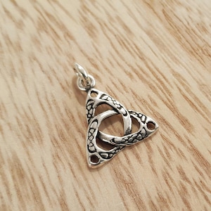 Op de afbeelding: Een zilverkleurige hanger in de vorm van een Keltische knoop, ook wel triquetra genoemd. De hanger heeft een textuurontwerp en een kleine lus aan de bovenkant voor een ketting. De hanger wordt gepresenteerd op een houten oppervlak.