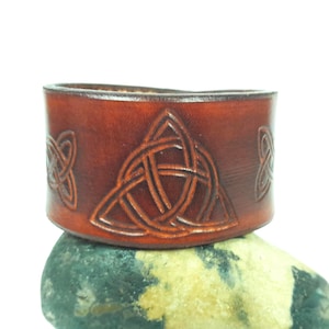 May include: A brown leather cuff bracelet with Celtic knot designs. The bracelet features a central triquetra knot and two smaller knots on either side. The leather has a rich, reddish-brown color and is set on a stone.