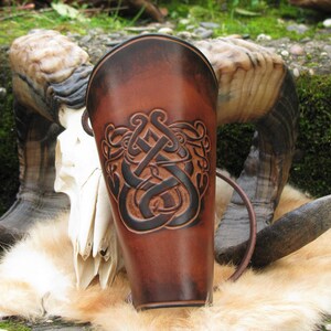Könnte beinhalten: Ein brauner Lederarmschutz mit einem keltischen Knotenmuster. Der zylindrische Armschutz hat ein dunkelbraunes geprägtes Muster und einen Lederriemen. Der Hintergrund umfasst einen Schädel und Tierfell.