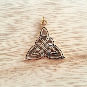 May include: A bronze-colored Celtic Triquetra pendant with a gold-colored loop for a chain. The pendant features an intricate interwoven design, with three interconnected arcs forming a triangular shape. The pendant is displayed on a light brown wooden surface.