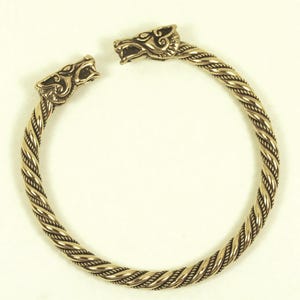 Könnte beinhalten: Ein goldfarbenes, offenes Armband mit einem gedrehten Seildesign. Das Armband hat detaillierte Wolfskopf-Terminals. Das Armband ist ein von Wikingern inspiriertes Schmuckstück.