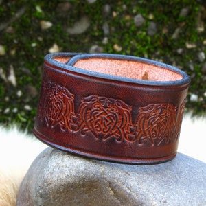 Könnte beinhalten: Ein braunes Lederarmband mit einem keltischen Knotendesign. Das Armband ist mit einem sich wiederholenden Muster aus verschlungenen Knoten geprägt.