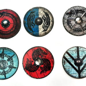 Puede incluir: Seis escudos vikingos redondos con varios diseños. Los escudos presentan una variedad de colores, incluyendo rojo, azul y gris, con intrincadas ilustraciones de dragones, cuervos y otros símbolos nórdicos. Cada escudo tiene un brocal central.