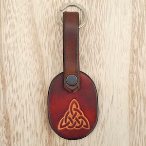May include: A brown leather keychain with a Celtic knot design. The keychain has a dark brown strap and a metal keyring. The oval-shaped pendant features an embossed Celtic knot in a lighter brown color, secured with a black snap.
