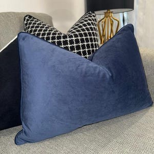 May include: A selection of decorative cushions in different designs. The front cushion is a solid, deep blue, velvet-like fabric. Behind it, a black and white geometric patterned cushion is visible. The cushions are arranged on a grey sofa.