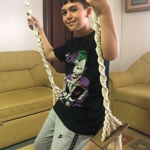 May include: A young person sits on a wooden swing with white rope supports. The person wears a black t-shirt with a Joker graphic and gray sweatpants. The swing is in a room with a yellow sofa.