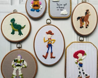 May include: A collection of seven embroidery hoops featuring needle-felted characters and text. Designs include a green dinosaur, a potato-shaped figure, a cowboy, a space ranger, a cowgirl, a horse, and the text You've got a friend in me.