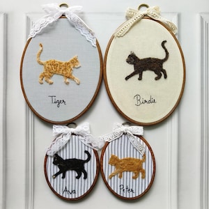 May include: Four oval-shaped cat embroidery hoops with wooden frames. Each features a felt cat silhouette and a name embroidered below. The backgrounds are light blue, cream, and striped fabric. White lace bows adorn the tops.