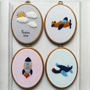 May include: Four oval embroidery hoops with felt designs. One features a sun, clouds, and the name "Parker JOHN". Others depict an orange and blue airplane, a rocket, and a blue and white airplane. Each is set in a wooden frame.