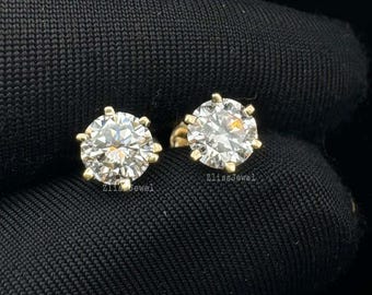 Puede incluir: Un par de pendientes de diamantes. Cada pendiente presenta un gran diamante redondo con talla brillante engastado en una montura dorada de seis puntas. Los pendientes se muestran sobre un fondo oscuro, resaltando su brillo.
