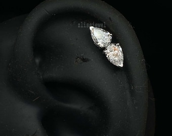 May include: A close-up of a black ear with two pear-shaped, clear gemstones. The gemstones are set in a silver or white metal and are positioned close together. The background is solid black.