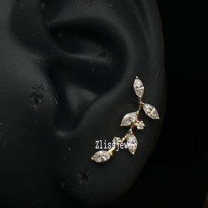 May include: A gold ear cuff with a leaf design, adorned with clear gemstones. The ear cuff is displayed on a black ear model. The text "Zlissjewel" is visible on the ear cuff.