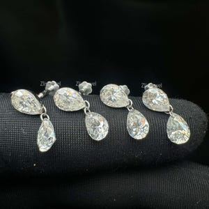 Puede incluir: Cuatro pares de pendientes colgantes de diamantes. Cada pendiente presenta un diamante en forma de pera con un diamante colgante en forma de pera. Los pendientes est&aacute;n engarzados en un metal plateado y se muestran sobre una superficie negra.