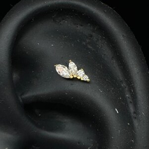 May include: A gold-colored ear piercing stud featuring three clear, marquise-cut gemstones and smaller round gemstones. The stud is set against a dark background, highlighting the jewelry's sparkle and design.