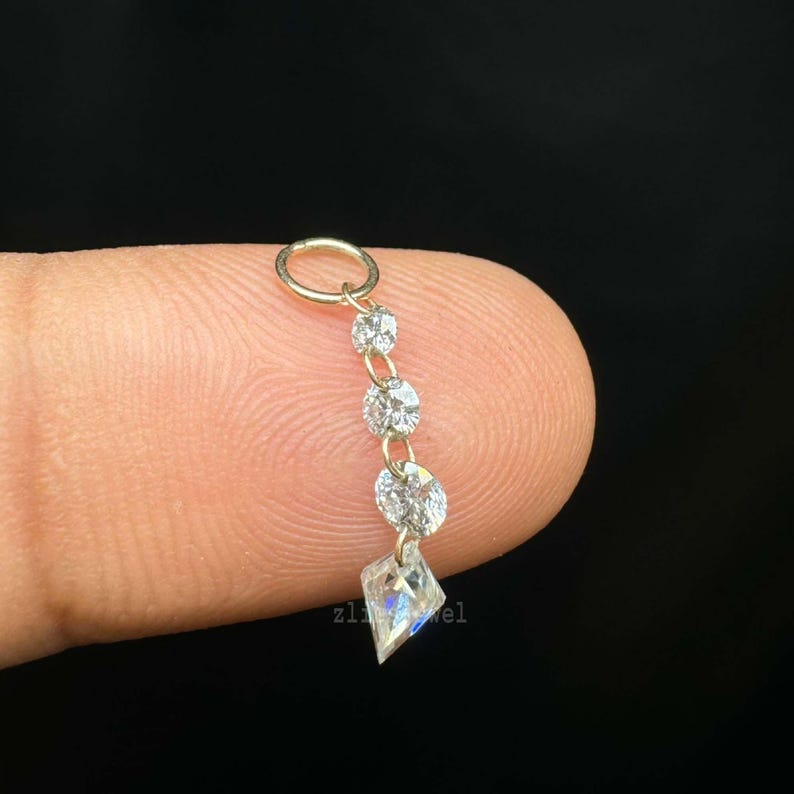 Puede incluir: Un peque&ntilde;o pendiente colgante con un aro dorado y una cadena de tres gemas redondas y transparentes, y una gema en forma de diamante en el extremo. La palabra "zl jewel" es visible.