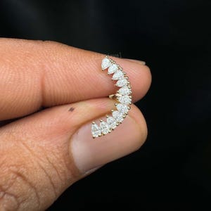 May include: A curved, gold-toned earring featuring a series of pear-shaped, clear gemstones. The gemstones are arranged in a crescent shape, creating a sparkling, elegant design. The earring is held against a dark background.