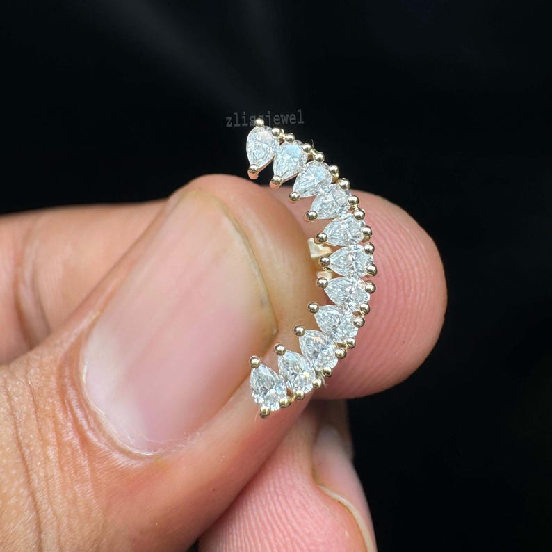 May include: A curved, gold-toned ear cuff featuring a row of pear-shaped, clear gemstones. The gemstones are set closely together, creating a sparkling, elegant design. The jewelry is held against a dark background.