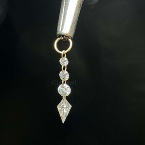 Puede incluir: Un pendiente dorado con un dise&ntilde;o colgante. El pendiente presenta tres gemas redondas y transparentes y una gema en forma de diamante en la parte inferior. El pendiente est&aacute; unido a una pieza de metal plateado. El fondo es negro.