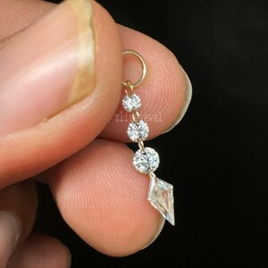 Puede incluir: Un aro de nariz dorado con un dise&ntilde;o colgante. El dise&ntilde;o presenta tres gemas redondas y transparentes y una sola gema transparente en forma de diamante. El texto "zlisjewel" es visible.