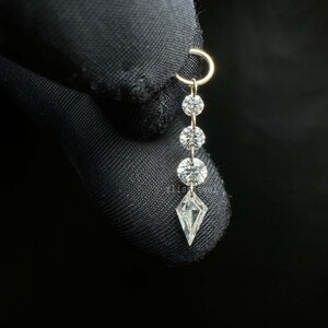 Puede incluir: Un pendiente dorado con cuatro piedras preciosas transparentes. La piedra superior est&aacute; unida a un aro circular, seguida de tres piedras redondas y una piedra en forma de cometa en la parte inferior. Fondo negro.