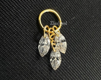 Puede incluir: Un aro de nariz dorado con tres piedras preciosas claras, de talla marquesa, colgando de un anillo circular. La joya está colocada sobre un fondo negro texturizado. El texto "ZlissJewel" es visible en el anillo.