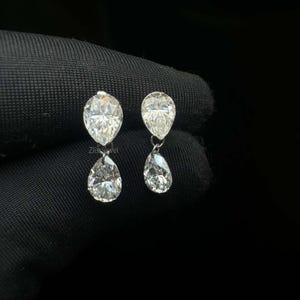 Puede incluir: Un par de pendientes colgantes de diamantes. Cada pendiente presenta un diamante en forma de pera suspendido debajo de un diamante en forma de pera m&aacute;s peque&ntilde;o. Los diamantes brillan sobre un fondo oscuro, mostrando su claridad y brillo. Los pendientes son un dise&ntilde;o de joyer&iacute;a cl&aacute;sico.