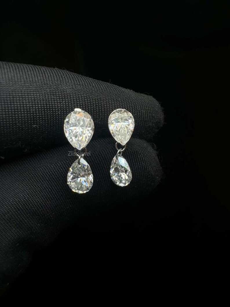 Puede incluir: Un par de pendientes colgantes de diamantes. Cada pendiente presenta un diamante en forma de pera suspendido debajo de un diamante en forma de pera m&aacute;s peque&ntilde;o. Los diamantes brillan sobre un fondo oscuro, mostrando su claridad y brillo. Los pendientes son un dise&ntilde;o de joyer&iacute;a cl&aacute;sico.