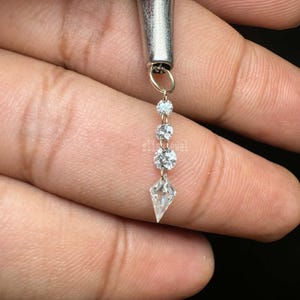 Puede incluir: Un pendiente plateado con un dise&ntilde;o colgante. El pendiente presenta tres piedras preciosas redondas y transparentes y una piedra preciosa en forma de cometa en la parte inferior. El pendiente est&aacute; sujeto por un cierre plateado. La palabra "zlisjewel" es visible.