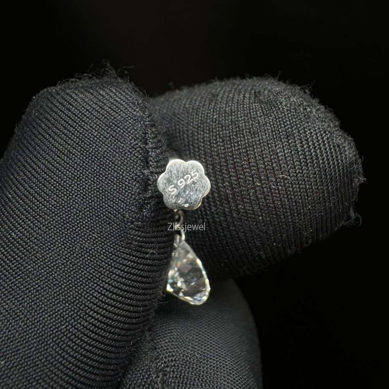 Puede incluir: Un pendiente de plata con un perno en forma de flor marcado "S925" y una piedra preciosa transparente en forma de l&aacute;grima. El pendiente se sostiene sobre un fondo negro. El texto "Zlissjewel" est&aacute; debajo del perno.