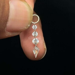 Puede incluir: Un delicado pendiente con un peque&ntilde;o aro y un dise&ntilde;o colgante. El pendiente presenta tres gemas redondas y transparentes y una gema transparente en forma de diamante en la parte inferior. La palabra "zlisejewel" es visible.