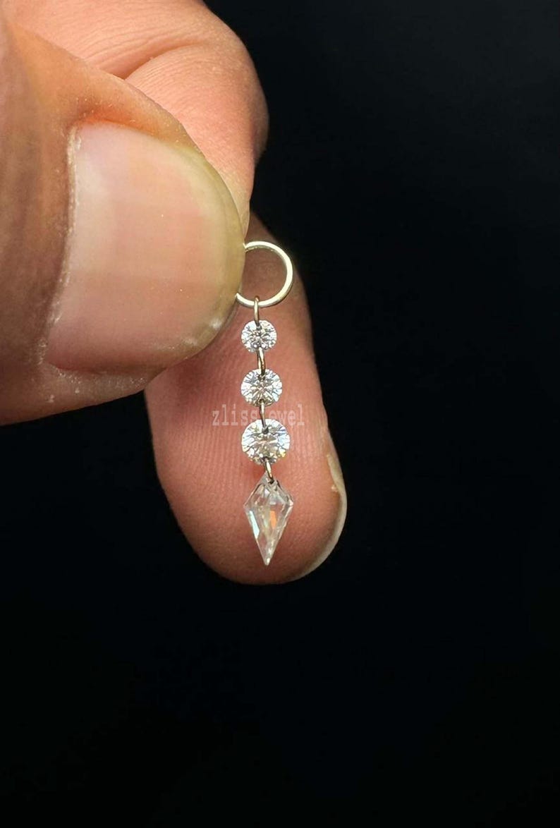 Puede incluir: Un delicado pendiente con un peque&ntilde;o aro y un dise&ntilde;o colgante. El pendiente presenta tres gemas redondas y transparentes y una gema transparente en forma de diamante en la parte inferior. La palabra "zlisejewel" es visible.